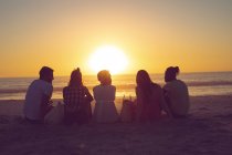 Rückansicht diverser Freunde, die bei Sonnenuntergang zusammen am Strand sitzen — Stockfoto