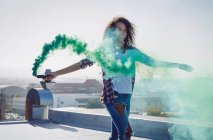 Front view of a young African-American woman wearing a denim vest holding a smoke maker producing green smoke on a rooftop with sunlight — Stock Photo