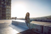 Frontansicht einer jungen afrikanisch-amerikanischen Frau in Lederjacke, die von der Kamera wegschaut, während sie auf einem Dach mit Blick auf ein Gebäude und das Sonnenlicht sitzt — Stockfoto