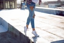 Lower half of a woman wearing ripped jeans and white sneakers on a rooftop with sunlight — Stock Photo