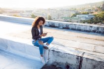 Vista lateral de uma jovem afro-americana usando uma jaqueta de couro enquanto se senta e usa um telefone celular em um telhado — Fotografia de Stock