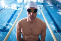 Front view of a male Caucasian swimmer wearing a white swimming cap and goggles standing by an olympic sized pool inside a stadium — Stock Photo