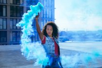 Front view of a young African-American woman wearing a denim vest holding a smoke maker producing blue  smoke on a rooftop with a view of a building and sunlight — Stock Photo