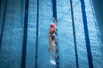 Vista ad alto angolo di una donna caucasica che indossa un cappello da bagno rosa e occhiali che fanno un ictus stile libero in una piscina — Foto stock
