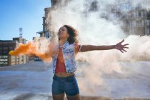 Vorderansicht einer jungen afrikanisch-amerikanischen Frau, die eine Jeansweste mit ausgestreckten Armen trägt und eine Rauchmaschine hält, die orangefarbenen Rauch auf einem Dach mit Blick auf ein Gebäude und Sonnenlicht produziert — Stockfoto