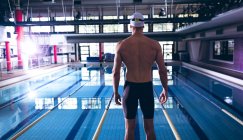 Vista trasera de un nadador caucásico masculino con una gorra blanca junto a una piscina olímpica dentro de un estadio - foto de stock