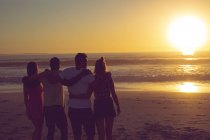 Rear view of diverse friends standing with arms around at beach during sunset — Stock Photo