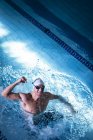 Vue en angle élevé d'un homme nageur caucasien portant un bonnet de bain blanc et des lunettes avec un poing levé souriant tout en se tenant dans la piscine — Photo de stock