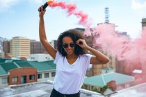 Vista frontal de uma jovem afro-americana vestindo uma camisa branca e óculos sorrindo enquanto segura um fabricante de fumaça produzindo fumaça vermelha em um telhado com vista para os edifícios — Fotografia de Stock