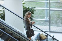 Вид збоку ділової жінки за допомогою мобільного телефону на ескалаторі в сучасному офісному корпусі — стокове фото