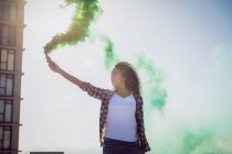 Front view of a young African-American woman wearing a plaid jacket holding a smoke maker producing green smoke on a rooftop with a view of a building and sunlight — Stock Photo