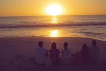 Вид сзади на друзей, сидящих вместе на пляже во время заката — стоковое фото