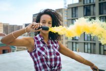 Vista frontal de una joven afroamericana vestida con una tapa a cuadros sosteniendo una máquina de humo que produce humo amarillo en una azotea con vista a edificios - foto de stock
