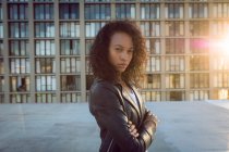 Vista laterale di una giovane donna afro-americana che indossa una giacca di pelle guardando attentamente la fotocamera con le braccia incrociate mentre si trova su un tetto con vista su un edificio e sul tramonto — Foto stock