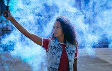 Frontansicht einer jungen afrikanisch-amerikanischen Frau in einer Jeansweste, die eine Rauchmaschine hält, die blauen Rauch produziert — Stockfoto
