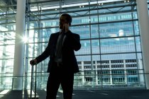 Frontansicht eines Geschäftsmannes mit Rollkoffer im Gespräch mit dem Handy im modernen Bürogebäude — Stockfoto