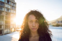 Primo piano di una giovane donna afro-americana che indossa una giacca di pelle guardando attentamente la telecamera mentre si trova su un tetto con vista su un edificio e sul tramonto — Foto stock