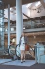 Vue de face d'une femme d'affaires marchant près d'un escalier roulant dans un immeuble de bureaux moderne — Photo de stock