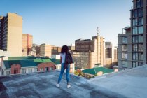 Back view of a young African-American woman with a leather jacket over shoulder standing on a rooftop with a view of buildings — Stock Photo
