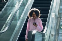 Vista frontal da empresária afro-americana olhando para o telefone celular enquanto usa escadas rolantes no escritório moderno — Fotografia de Stock