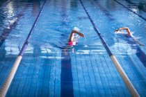 Vista frontal de una joven afroamericana y caucásica haciendo un derrame cerebral estilo libre en la piscina mientras que el nadador con gorra rosa lleva - foto de stock