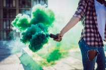 Mid section of a woman wearing a plaid jacket holding a smoke maker producing green smoke on a rooftop with a view of a building and sunlight — Stock Photo