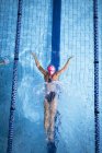 Hochwinkelaufnahme einer kaukasischen Frau mit pinkfarbener Badekappe und Brille, die in einem Schwimmbad einen Schmetterlingsschlag macht — Stockfoto