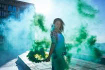 Vista frontal de una joven afroamericana con una chaqueta a cuadros sosteniendo una máquina de humo que produce humo verde en una azotea con vistas a un edificio y la luz del sol - foto de stock