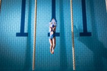 Vista de ángulo alto de una mujer afroamericana con un traje de baño y una gorra de natación azul buceando en la piscina - foto de stock