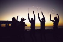 Frontansicht der Silhouette diverser Freunde, die in der Abenddämmerung mit Wunderkerzen am Strand spielen — Stockfoto