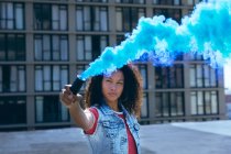 Front view of a young African-American woman wearing a denim vest holding a smoke maker producing blue smoke on a rooftop with a view of a building — Stock Photo