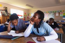Vista lateral de cerca de una joven colegiala africana apoyada en su escritorio, mirando hacia arriba y pensando mientras escribe en su cuaderno durante una lección
. - foto de stock