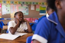 Vista frontal de un joven colegial africano sonriente sentado en su escritorio, apoyado y escuchando durante una lección en un aula de la escuela primaria del municipio, a su alrededor compañeros de clase también están sentados en sus escritorios y escuchando - foto de stock