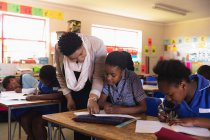 Visão frontal de uma professora de escola africana de meia-idade ajudando uma jovem estudante africana sentada em sua mesa durante uma aula em uma sala de aula da escola primária da cidade, ao lado dela e nos colegas de classe de fundo estão escrevendo em seus livros — Fotografia de Stock
