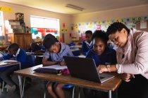 Nahaufnahme einer afrikanischen Lehrerin mittleren Alters, die einer jungen afrikanischen Schülerin hilft, während einer Unterrichtsstunde mit einem Laptop an ihrem Schreibtisch zu sitzen. — Stockfoto