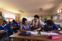 Vue de face d'une enseignante africaine d'âge moyen aidant une jeune écolière africaine assise à son bureau pendant une leçon dans une classe de l'école élémentaire d'un canton, à côté d'elle et en arrière-plan, des camarades de classe écrivent dans leurs livres — Photo de stock