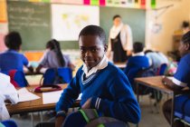 Vista lateral de cerca de un joven colegial africano sentado en su escritorio y dando la vuelta, mirando a la cámara y sonriendo durante una lección en un aula de la escuela primaria del municipio
. - foto de stock