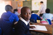 Vue de côté gros plan d'un jeune écolier africain assis à son bureau et se retournant, regardant vers la caméra pendant une leçon dans une classe de l'école élémentaire d'un canton
. — Photo de stock