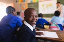Vista lateral de cerca de un joven colegial africano sentado en su escritorio y dando la vuelta, mirando a la cámara y sonriendo durante una lección en un aula de la escuela primaria del municipio . - foto de stock