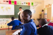 Vista lateral de cerca de una joven colegiala africana sentada en su escritorio y dando la vuelta, mirando a la cámara y sonriendo durante una lección en un aula de la escuela primaria del municipio
. - foto de stock
