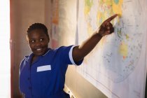 Вид спереди на молодую африканскую школьницу, стоящую перед классом, улыбающуюся и указывающую на карту во время урока в классе игрушечного корабля — стоковое фото
