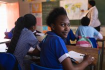 Seitenansicht eines jungen afrikanischen Schülers, der an seinem Schreibtisch sitzt und sich umdreht, in die Kamera blickt und während einer Schulstunde in einer Township-Grundschule lächelt. — Stockfoto