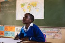 Вид сбоку на молодого африканского школьника, сидящего за столом и смотрящего вверх во время записи в записной книжке и внимательно слушающего во время урока в городском классе начальной школы — стоковое фото