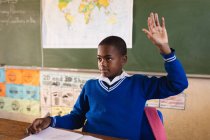 Vista frontal de perto de um jovem estudante africano sentado em uma mesa e levantando a mão para responder a uma pergunta durante uma aula em uma sala de aula da escola primária da cidade, em segundo plano é um mapa do mundo e do quadro-negro — Fotografia de Stock