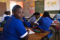 Vista laterale da vicino di un giovane scolaro africano seduto alla sua scrivania e che si gira, guarda la telecamera e sorride durante una lezione in una classe della scuola elementare cittadina . — Foto stock