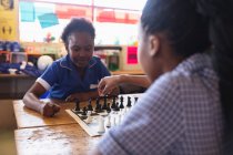 Over the shoulder view of two young African schoolgirls sitting at a desk playing chess during a break from lessons in a township elementary school classroom — Stock Photo