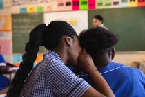 Vista trasera de cerca de dos jóvenes colegialas africanas sentadas en su escritorio susurrándose unas a otras durante una lección en un aula de la escuela primaria del municipio - foto de stock
