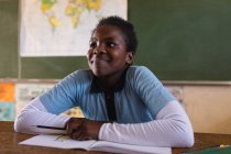 Vista frontal de cerca de una joven colegiala africana sentada en un escritorio sonriendo y escuchando atentamente durante una lección en un aula de la escuela primaria del municipio - foto de stock
