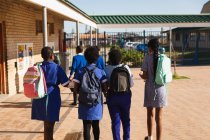 Rückansicht von vier jungen afrikanischen Schülerinnen, die mit ihren Taschen über den Schulhof einer Township-Grundschule laufen — Stockfoto