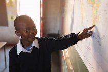 Front view close up of a young African schoolboy standing at the front of class smiling and pointing to a map during a lesson in a township elementary school classroom — Stock Photo